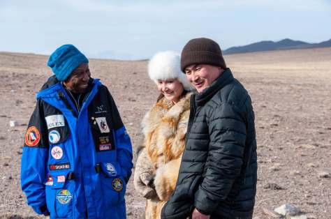 Barbara in a blue beanie and blue jacket, stands next to a Mongolian woman in a fur hat, coat, and falconers glove and a Mongolian man in a brown beanie and black coat. They are laughing together with a desert and mountain ridge behind them.